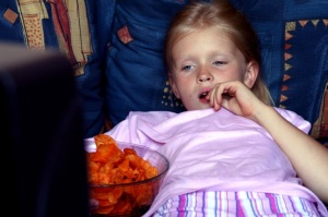 USA Elementary Age Girl Lying Down and Watching TV with Bowl of Carb Snacks Larger than Her Abdomen 