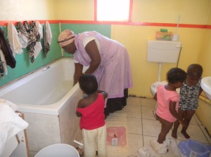 Woman cleaning the bathtub while 3 toddler age orpans watch.  The orphanage for AIDS victims has indoor plumbing.